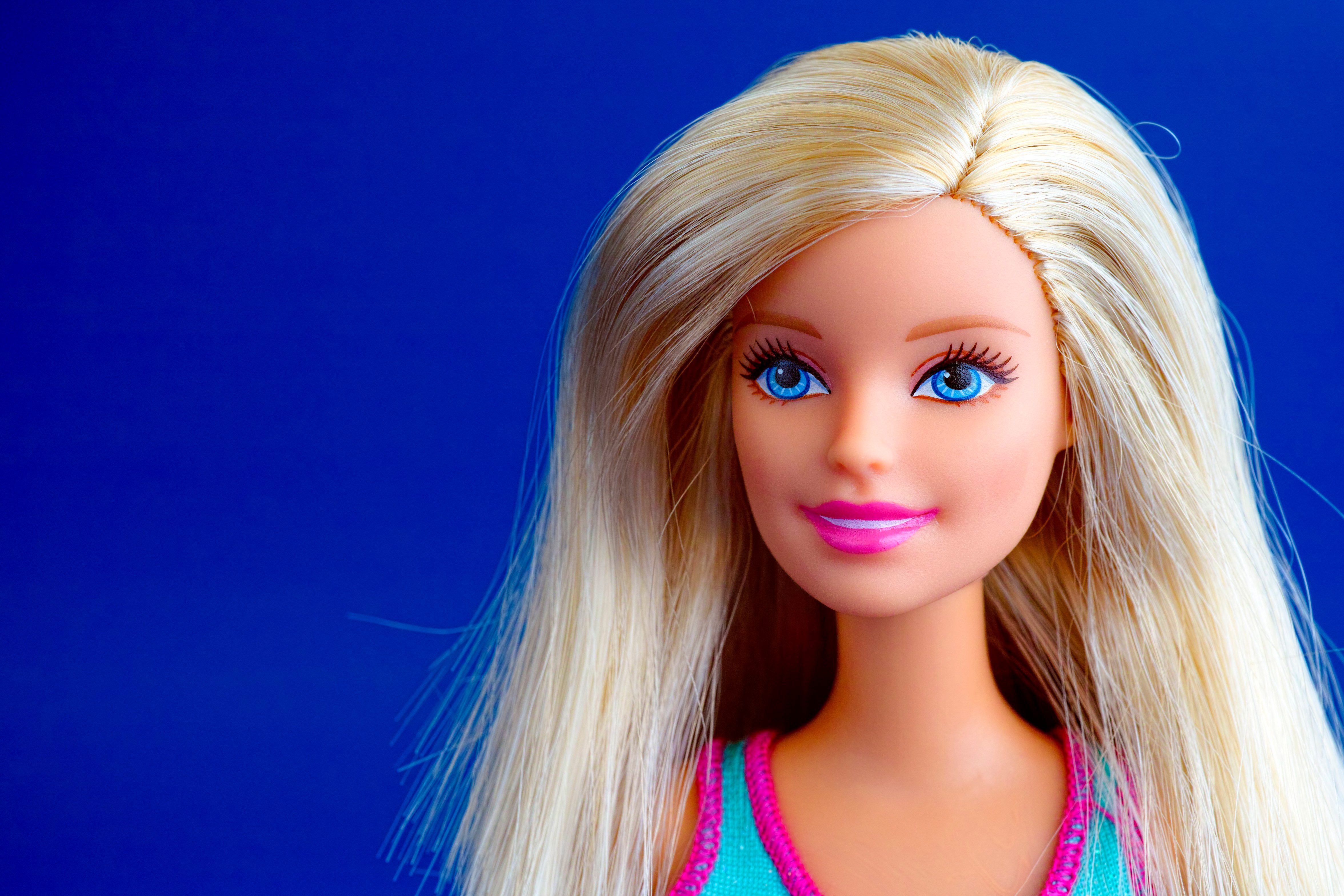 A blonde doll with bright blue eyes and pink lips smiles against a solid blue background.