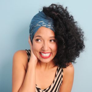 A woman, smiling and holding her face, wears a blue headwrap and striped top against a light blue background.
