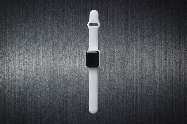 A white smartwatch lies flat on a dark, textured, metallic surface background, with its screen facing upwards.