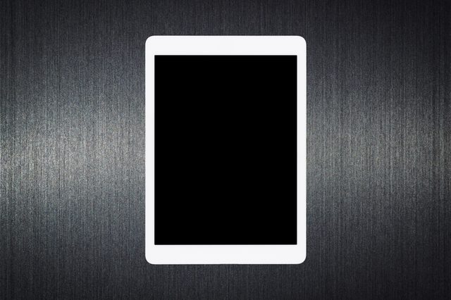A white tablet lies on a textured, dark surface, with its screen turned off, displaying a solid black display.