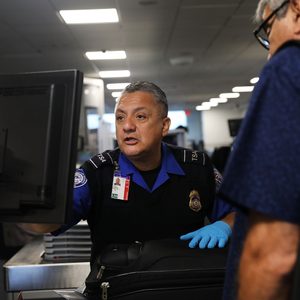 Security officer inspects luggage at an airport checkpoint, engaging with a traveler. Fluorescent lights illuminate the busy terminal scene.