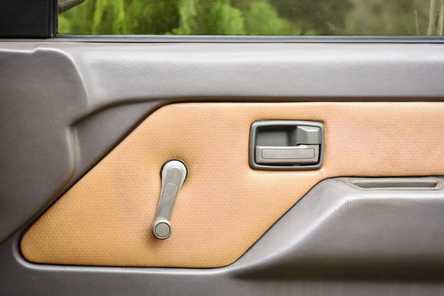 Car door panel features a manual window crank and handle, with beige upholstery and gray accents, positioned against a blurred greenery background.