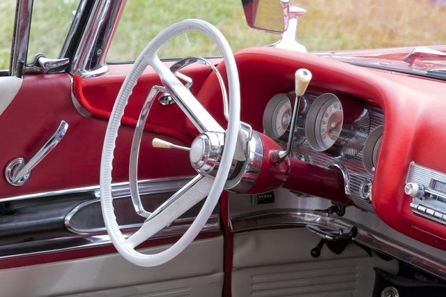 White steering wheel rests inside a classic car's red interior, featuring chrome details, gauges, and levers, with a grass field visible outside.