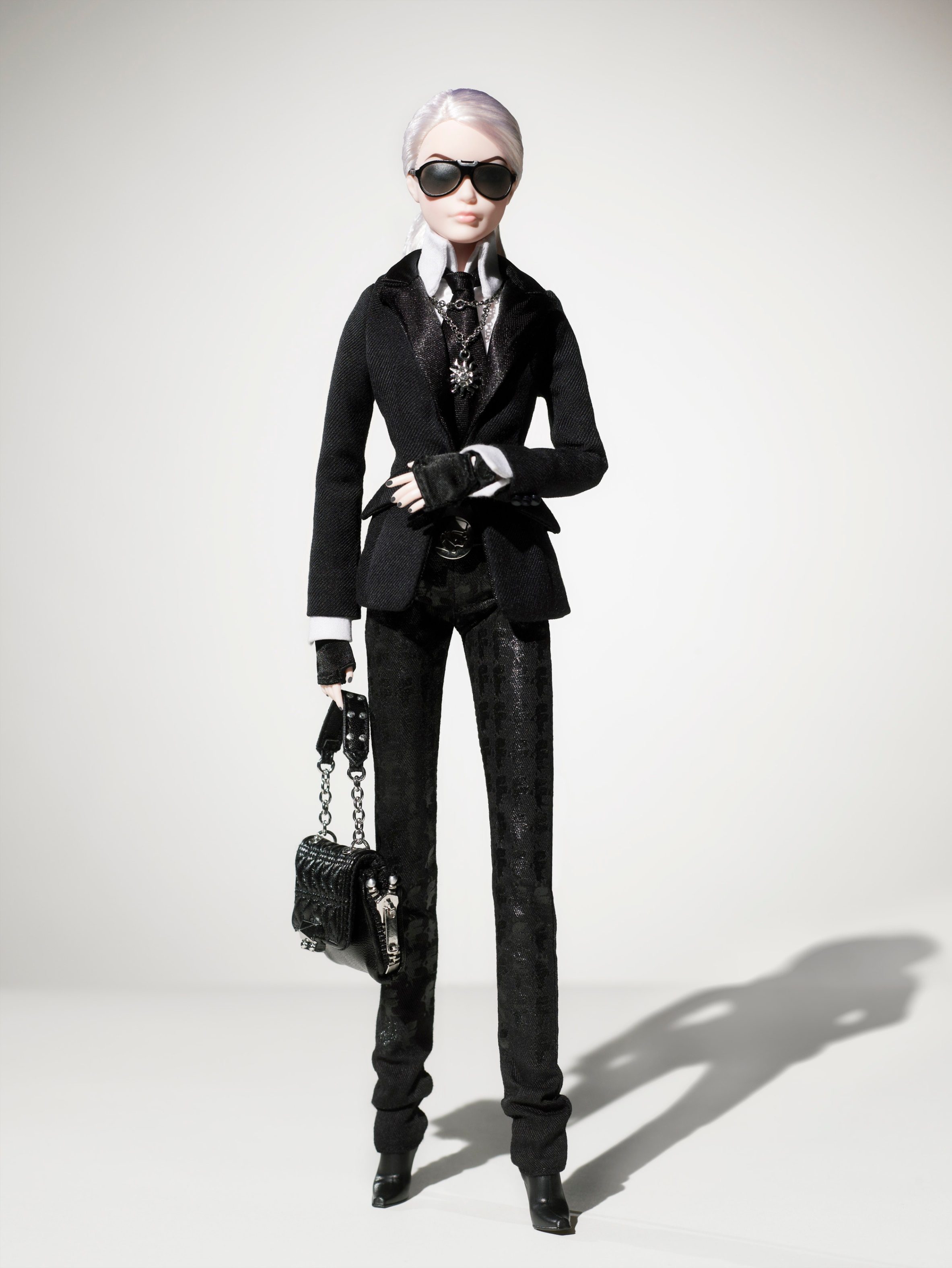 Doll stands confidently, wearing black suit, sunglasses, and holding a small handbag in a minimalist setting.