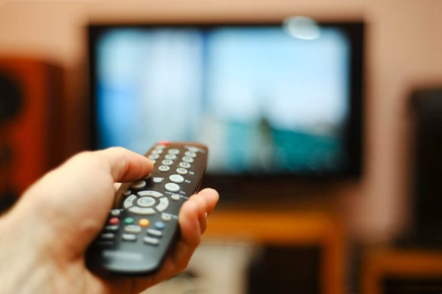 Hand holds remote, pointing at a blurry, active TV screen in a living room setting.