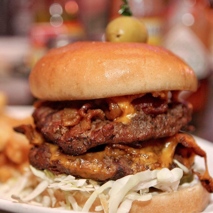 A double cheeseburger sits on a plate, featuring bacon, shredded lettuce, and a green olive on top, amidst a blurred restaurant setting.