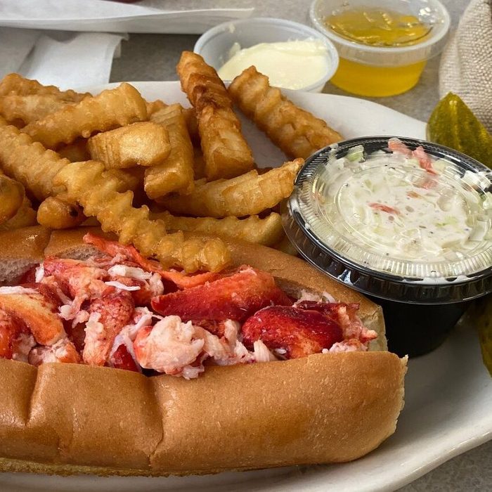 A lobster roll sits with crinkle-cut fries and coleslaw on a white plate, accompanied by a pickle slice and small sauce containers.