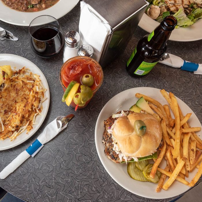 Burger with fries on a plate sits on a table, alongside a Bloody Mary, hash browns, and a beverage bottle, surrounded by other dishes.