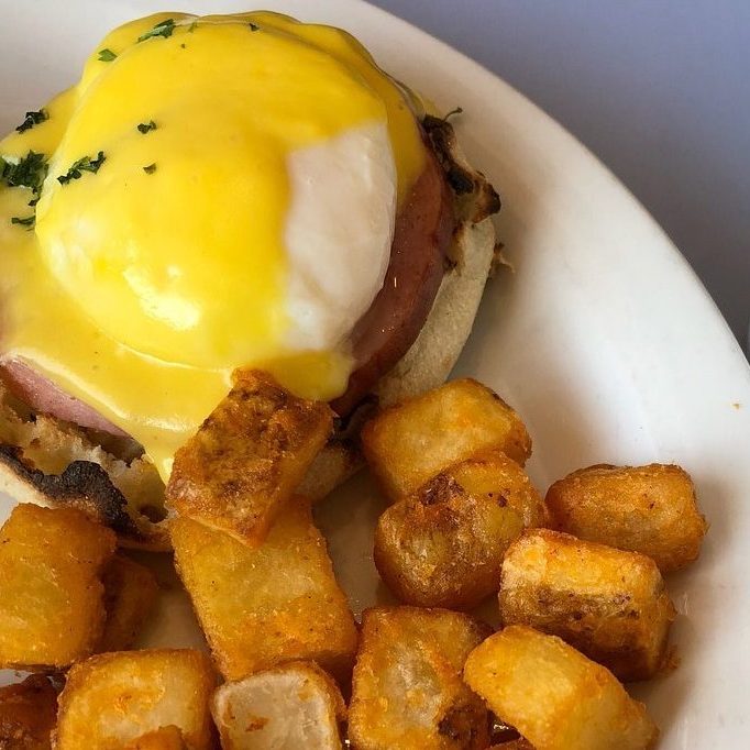 Eggs Benedict topped with hollandaise sauce sits on a plate, accompanied by golden-brown cubed potatoes.