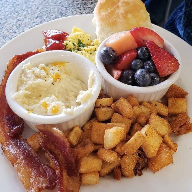 A breakfast plate features bacon, scrambled eggs, grits, diced potatoes, mixed fruit, and a biscuit, neatly arranged on a white surface.