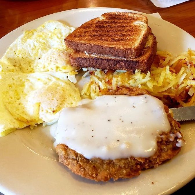 Breakfast plate holds sunny-side-up eggs, crispy toast, hash browns, and gravy-smothered chicken-fried steak. It's on a restaurant table.