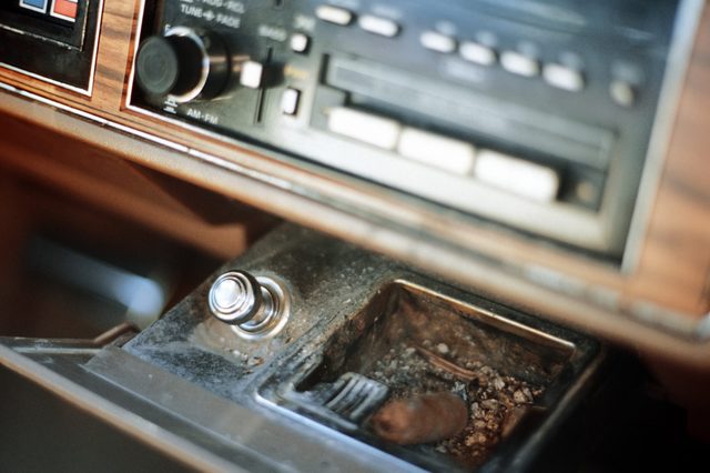 Car ashtray holds cigarette butt; surrounded by retro dashboard with radio and buttons, hinting at an older vehicle interior.