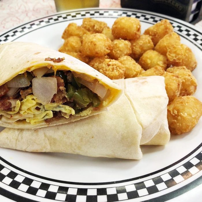 Burrito filled with eggs, bacon, and vegetables sits on a plate with a pile of golden tater tots in a diner setting.