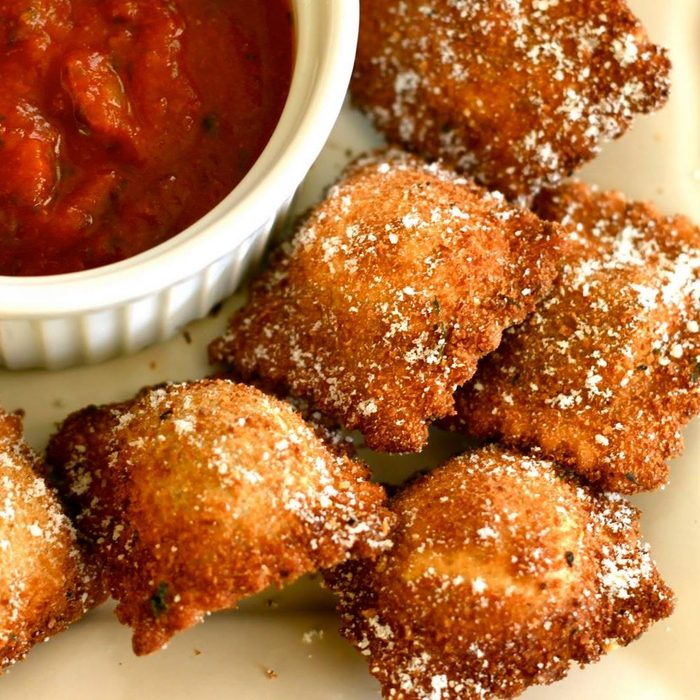 Crispy ravioli rest alongside a bowl of marinara sauce, sprinkled with grated cheese on a white plate.