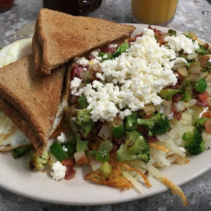 Toast and eggs sit beside a colorful vegetable hash topped with crumbled cheese on a white plate in a diner setting.