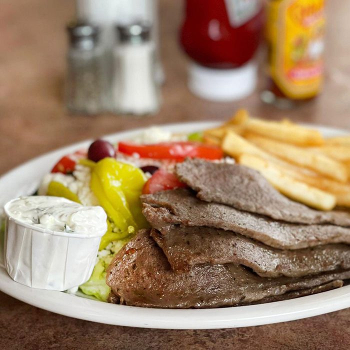A plate holds sliced meat, fresh salad, and fries, with tzatziki sauce, with salt, pepper, ketchup, and hot sauce bottles in the background.
