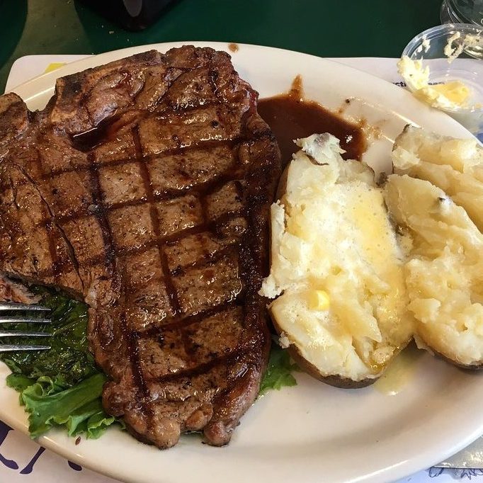 Grilled steak rests on lettuce with baked potato on a white plate, surrounded by green tablecloth, fork nearby.