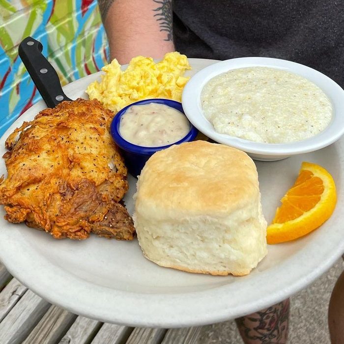 Plate holding fried chicken, biscuit, grits, scrambled eggs, gravy, and an orange slice; colorful mural visible in the background.