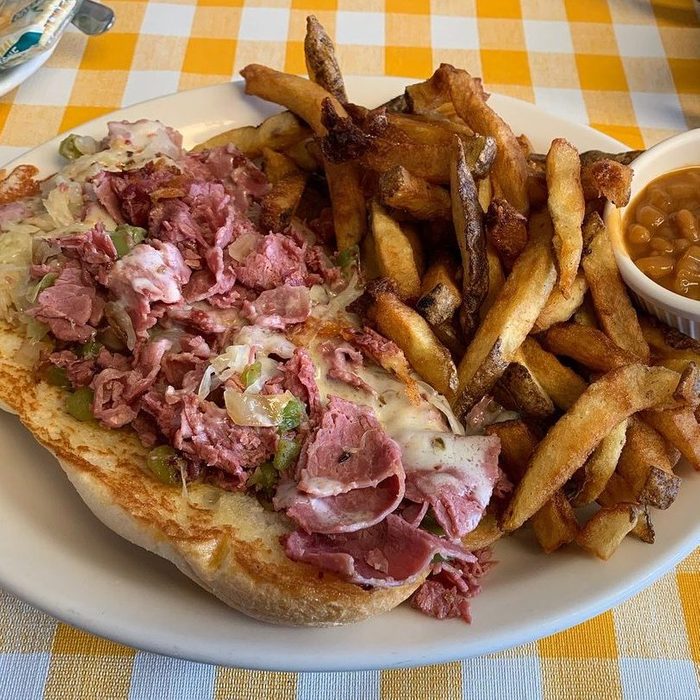 Sandwich filled with corned beef and melted cheese lies open, next to golden fries and a small cup of baked beans on a checkered tablecloth.