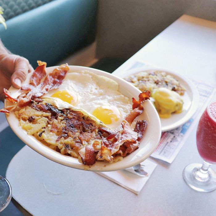 Plate holds bacon, eggs, and hash browns, served in a diner setting. A hand holds the plate over a table with more dishes, including a red drink.