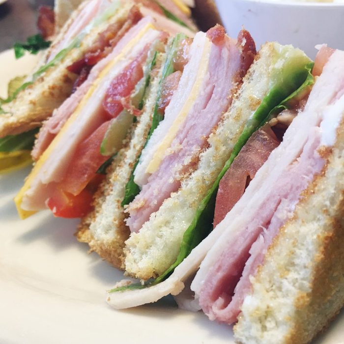 Sandwich slices stacked with turkey, bacon, lettuce, tomato, and cheese on toasted bread, placed on a plate in a dining setting.