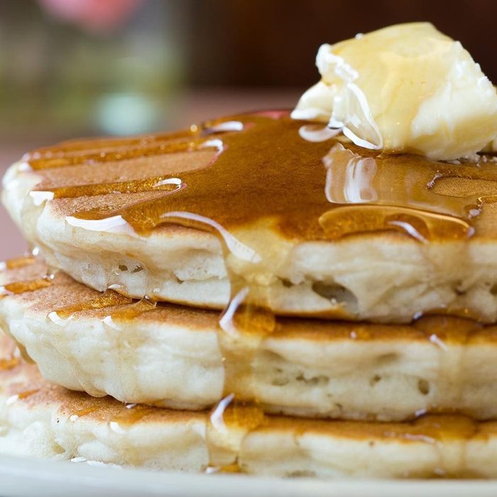 Pancakes stacked with syrup dripping topped with butter in a close-up view on a plate.