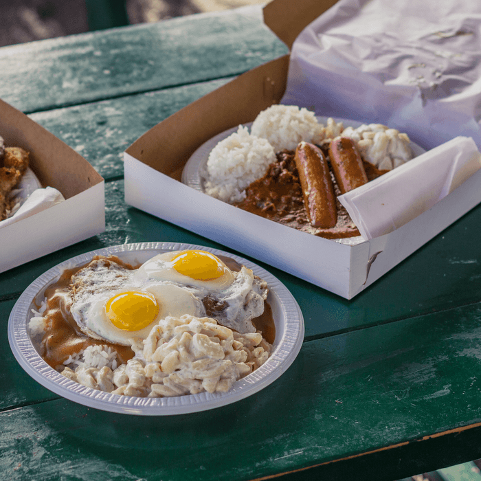Plates with eggs, macaroni, rice, and sausages sit on a green outdoor picnic table, surrounded by open cardboard takeout boxes.