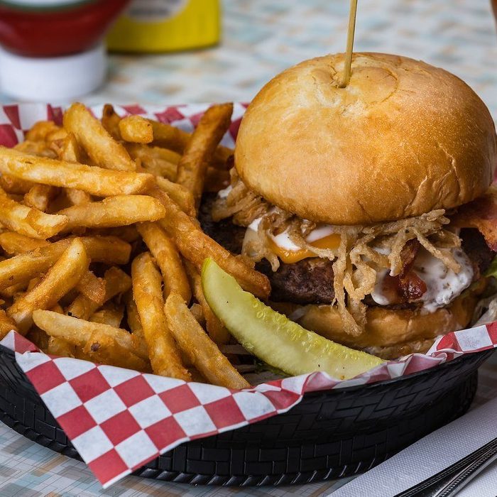 Burger topped with crispy onions sits on a basket; surrounded by golden fries, pickle slice, and red-checkered paper, in a diner setting.