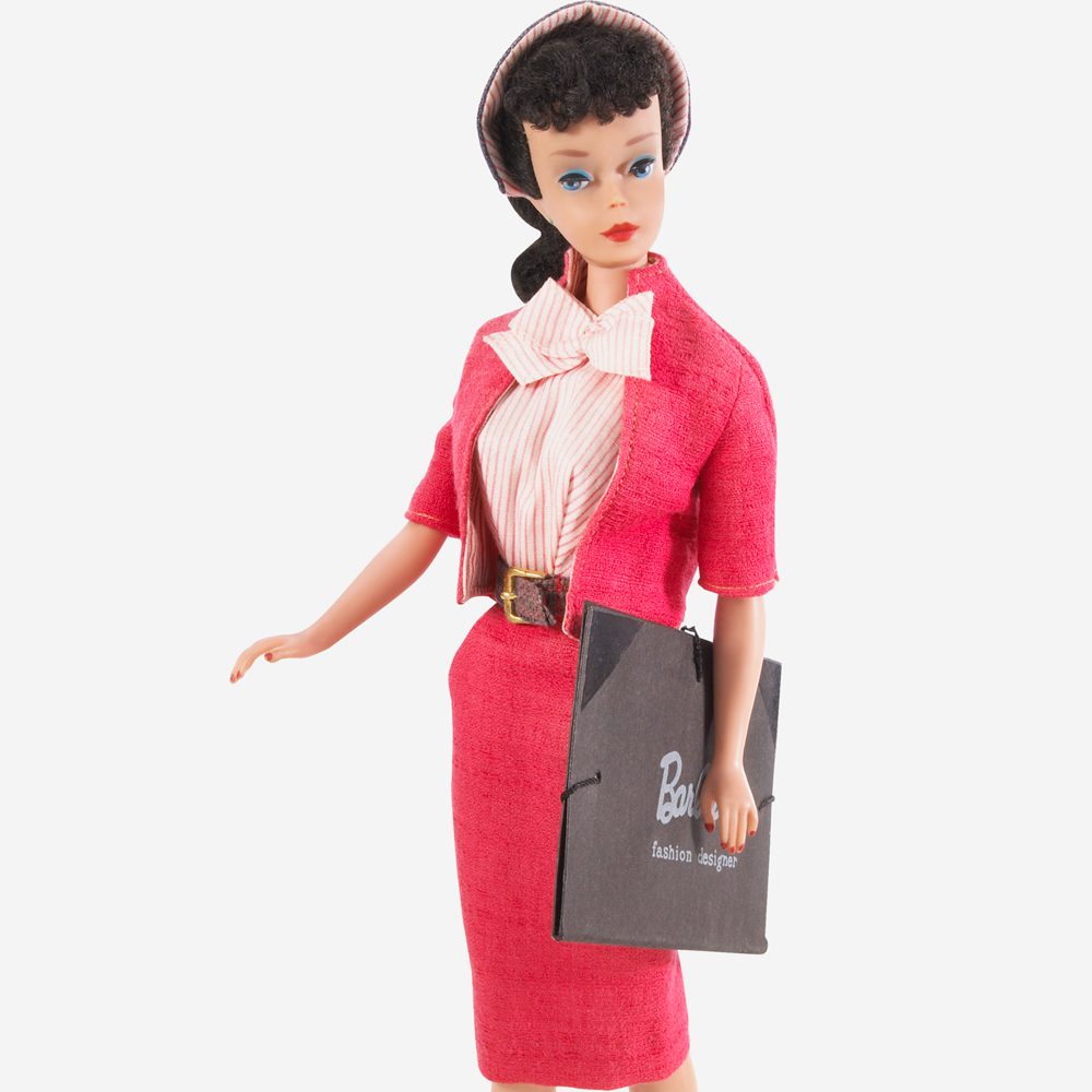 A doll holds a black shopping bag, dressed in a pink suit with a striped shirt, set against a plain white background.