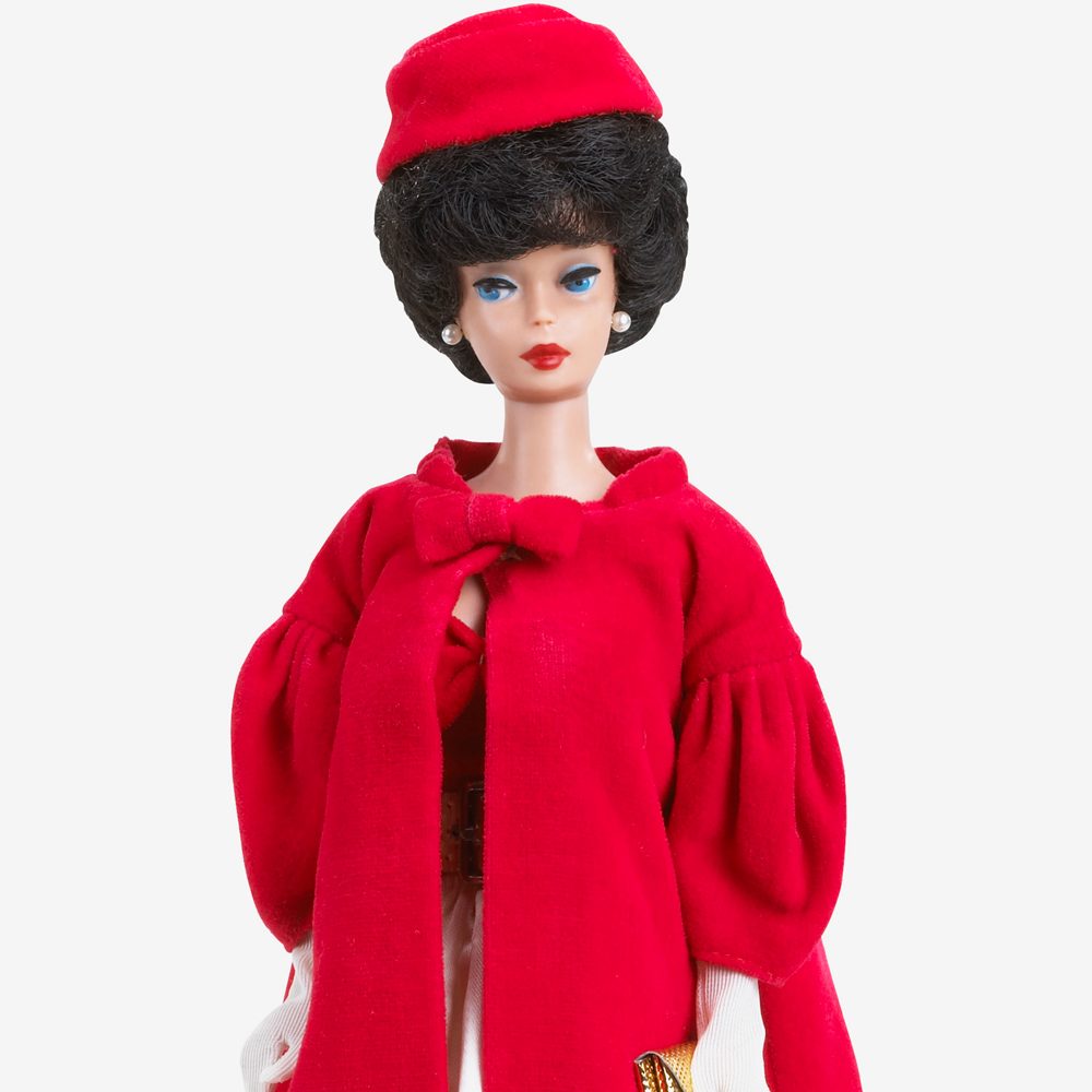 A doll wears a red coat and matching hat, with short dark hair, against a plain white background.