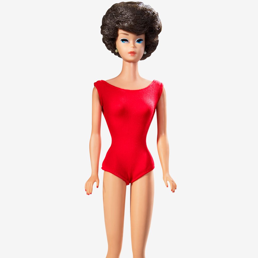 Doll stands, wearing a red one-piece swimsuit, with short dark hair, set against a plain white background.