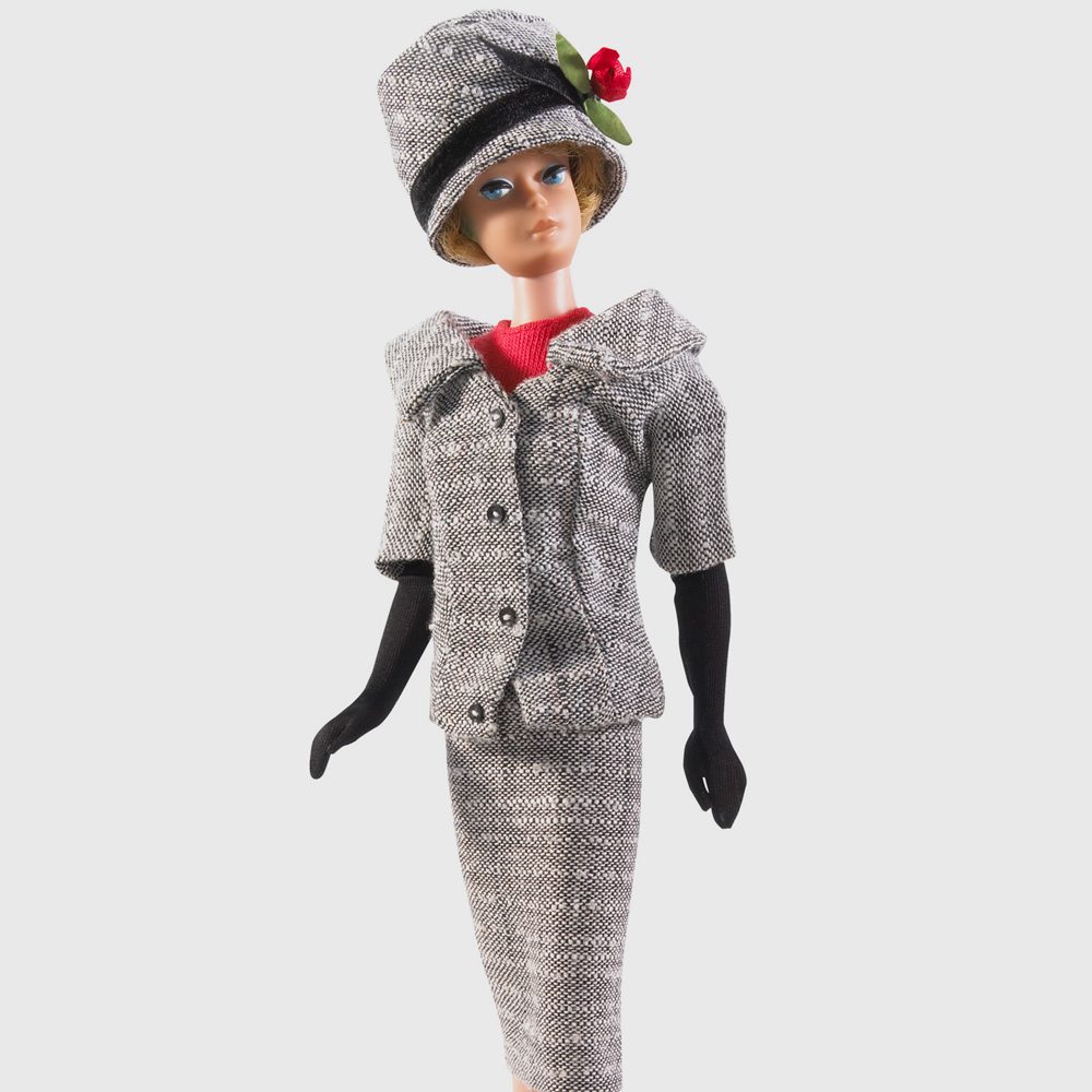 Doll wearing a gray tweed outfit and hat stands upright, adorned with black gloves and a red flower, against a plain background.