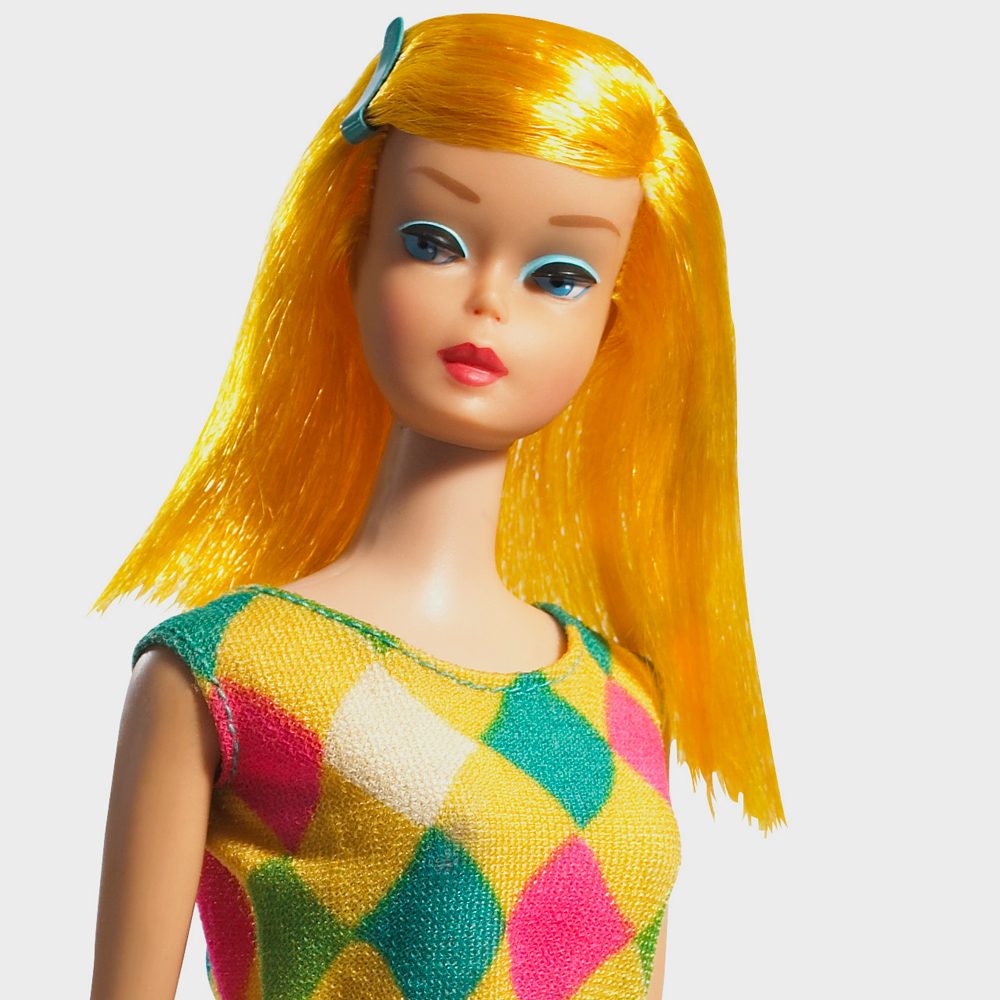 Doll stands, wearing a colorful checkered dress, with bright yellow hair styled straight and a blue hair clip, on a plain background.