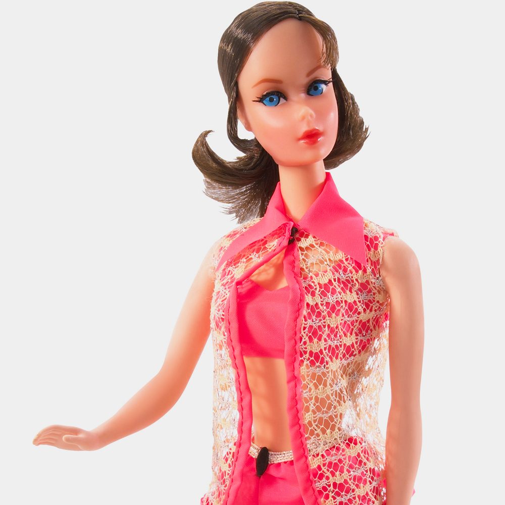 Doll poses in pink and lace outfit against a plain background, hair styled in a retro flip.