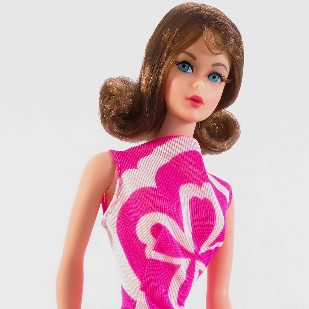Doll stands with a confident pose, wearing a pink and white patterned dress, set against a plain background.