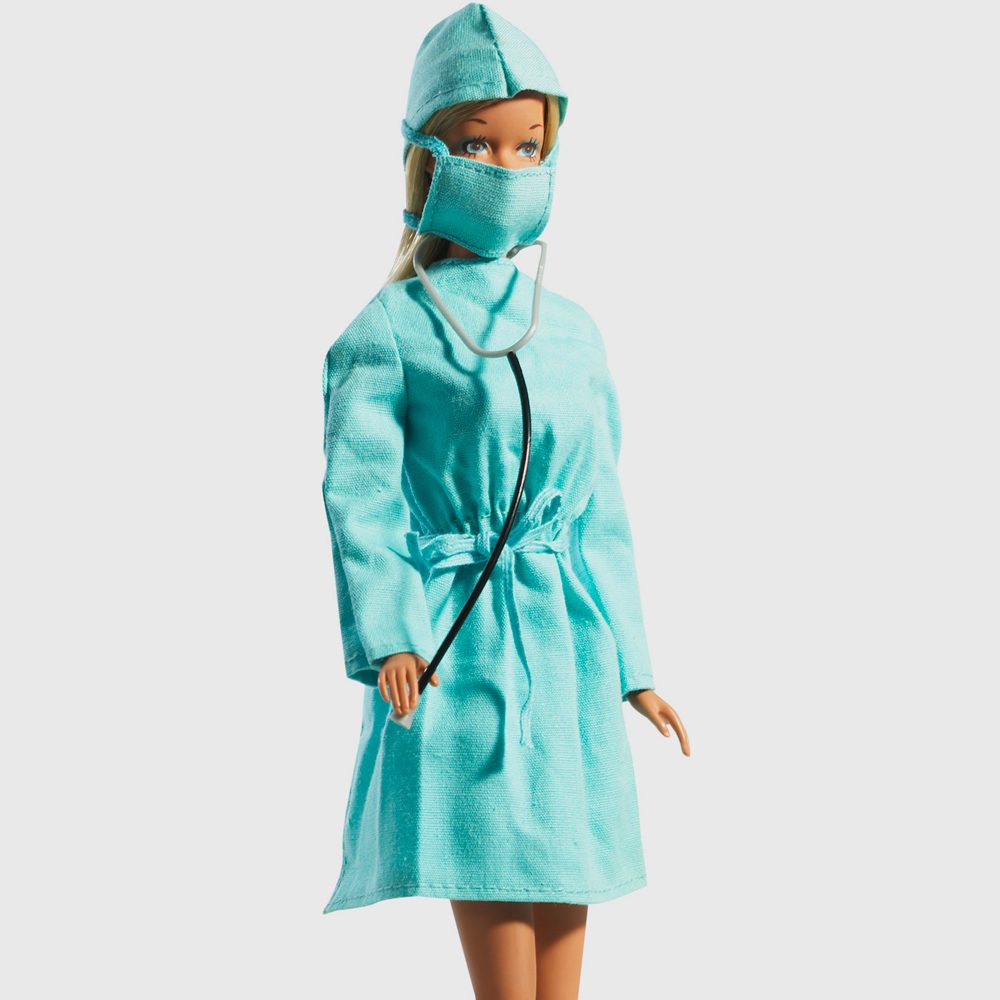 A doll wears a turquoise surgical outfit with a mask, holding a stethoscope against a plain white background.