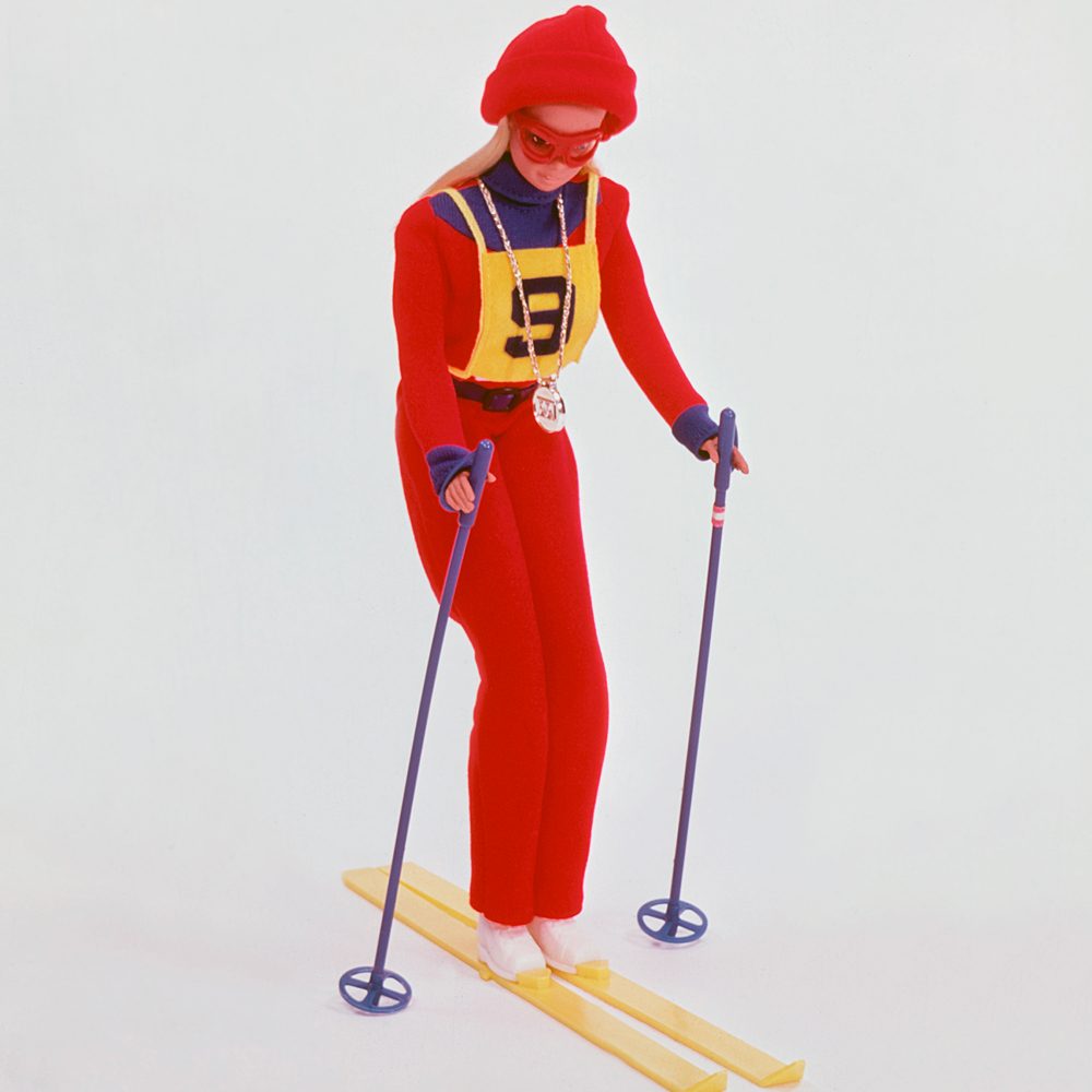 Doll skis downhill, wearing a red and yellow outfit with matching accessories, against a plain white background.