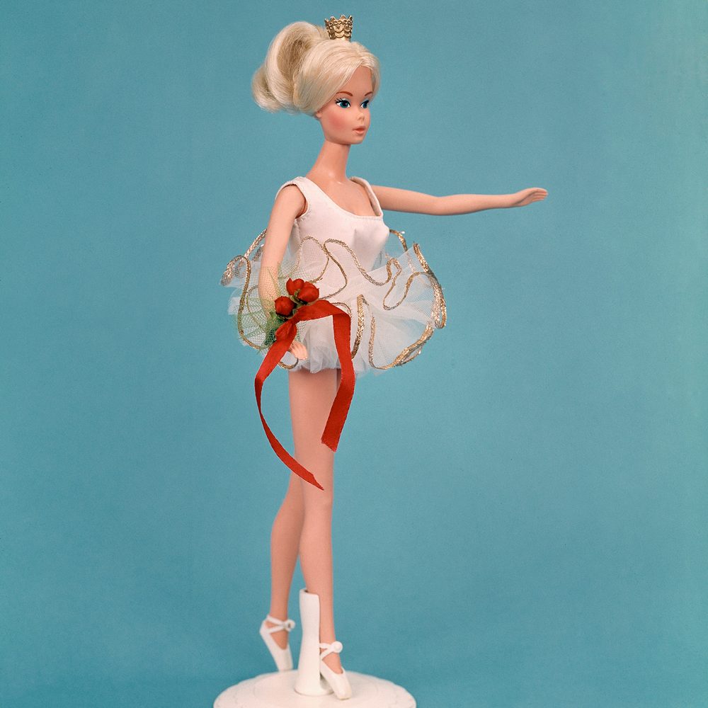 Doll dressed as ballerina stands on one leg, arm outstretched, wearing crown and white tutu with gold accents, against plain blue background.