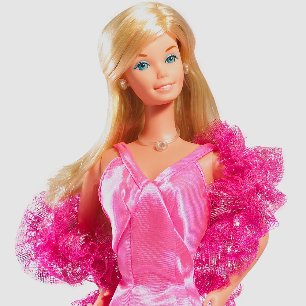 A doll stands wearing a shiny pink dress with ruffled trim, featuring long blonde hair and a string necklace on a white background.
