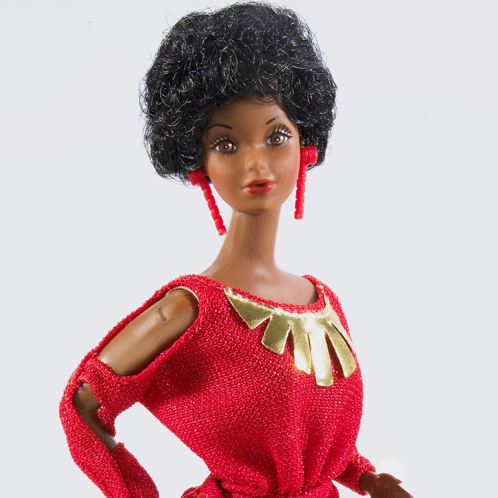 Doll wearing a red dress with gold accents poses confidently, sporting black curly hair and red earrings in a bright, plain background.