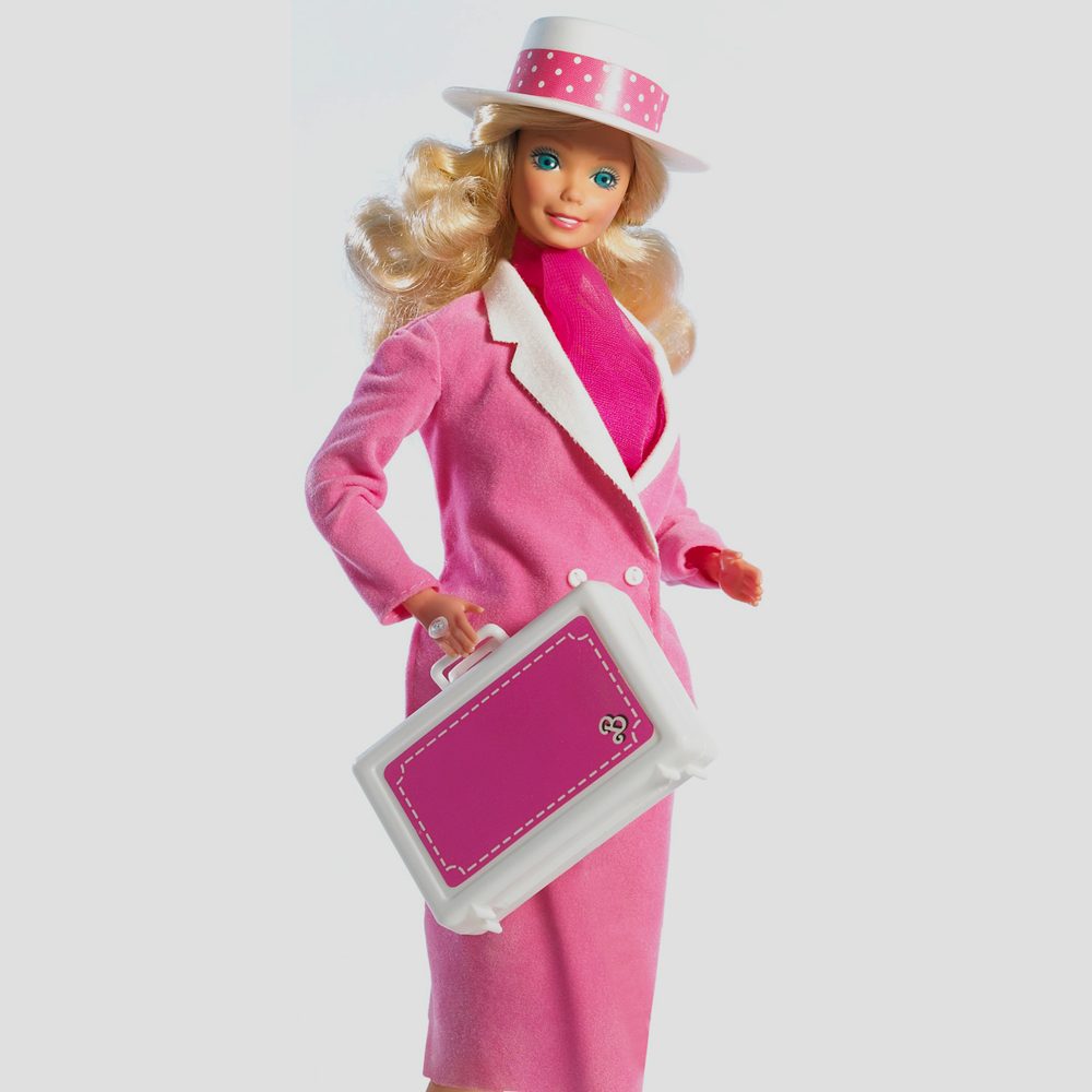 A doll holding a pink suitcase wears a pink suit and hat, posed against a plain white background.