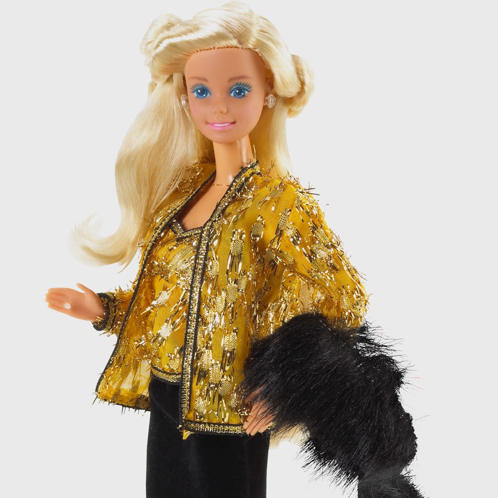 A doll poses wearing a gold sparkling outfit and black fur stole against a plain white background.