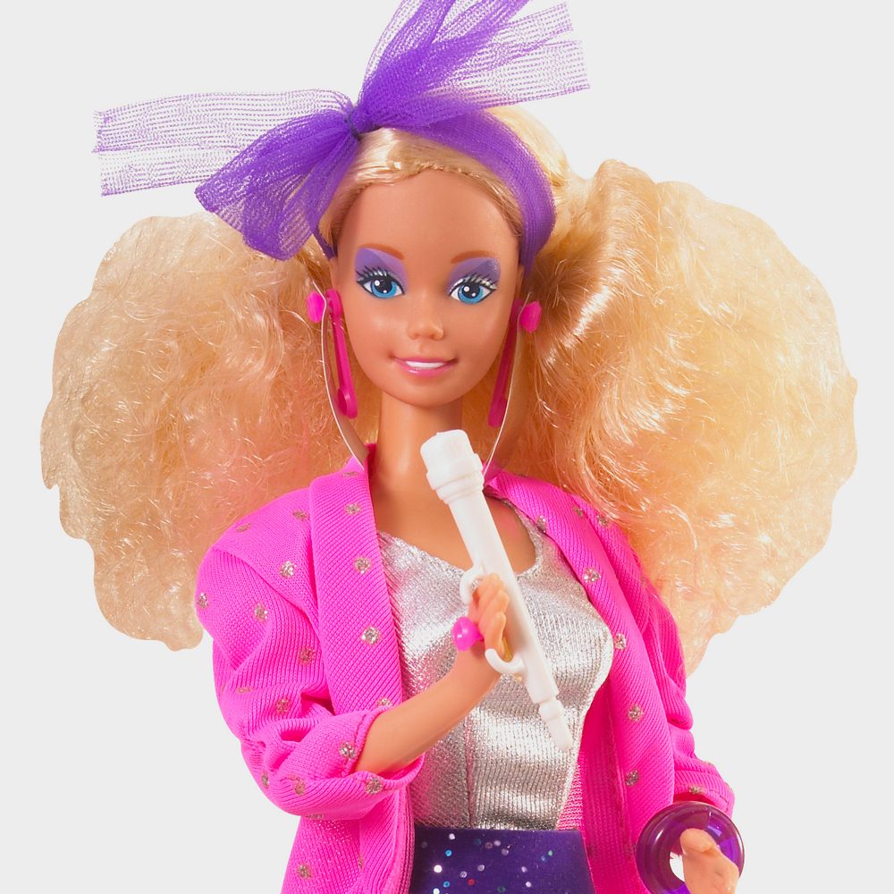 Doll holds a microphone, wearing a bright pink jacket and purple headband, standing against a plain background.