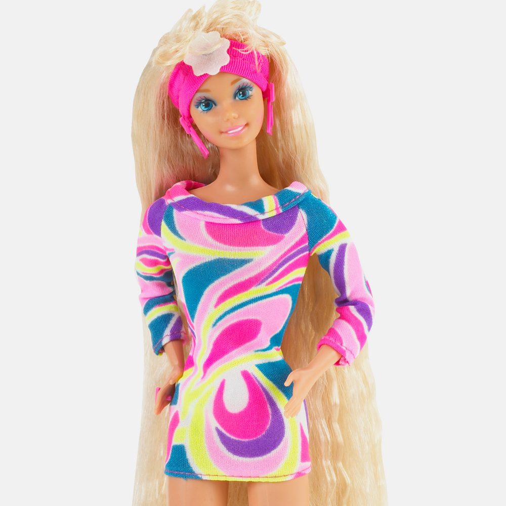 Doll wears colorful, swirl-patterned dress with a pink headband, standing confidently against a plain background.