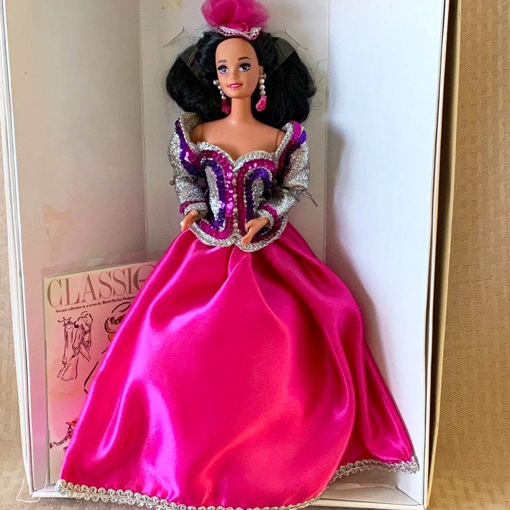 Doll wears sparkling, vibrant pink and silver dress, with dark hair, in a doll box. Inside, "CLASSICS" text visible on accompanying material.