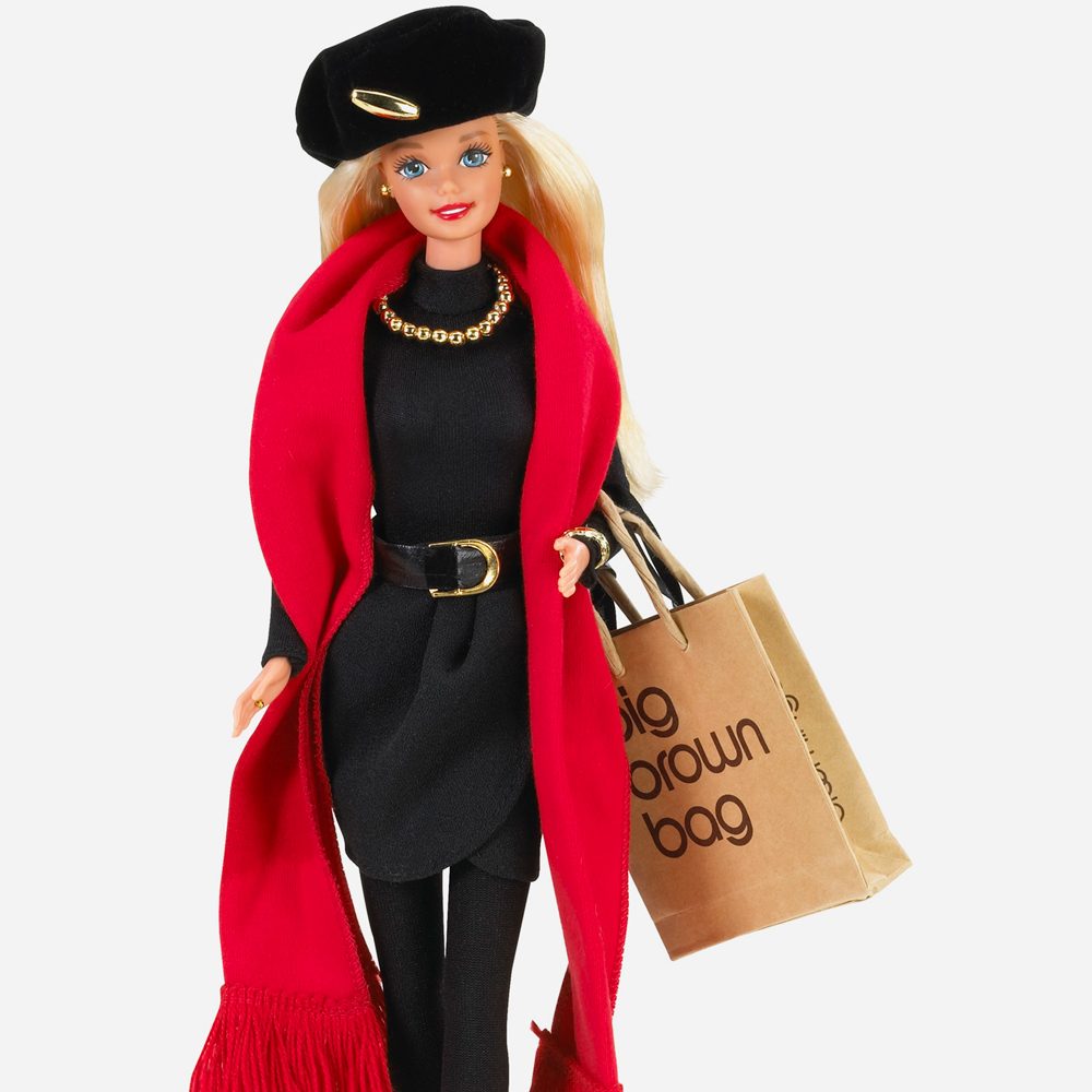 Doll wears black outfit, red scarf, holding shopping bag marked "big brown bag," against plain background.