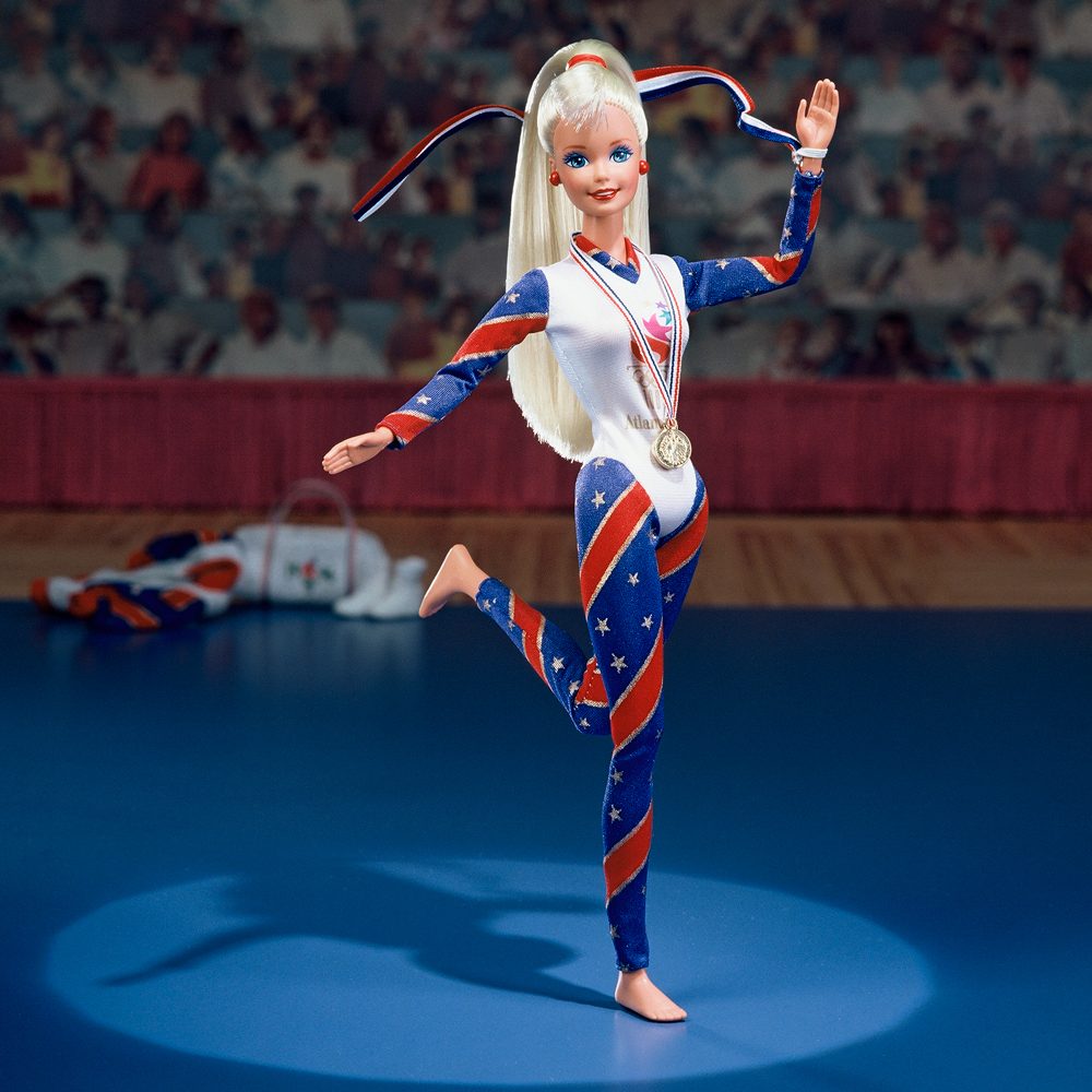 Doll poses proudly, wearing a patriotic-themed leotard with a medal, on a blue gym floor, surrounded by an audience backdrop.