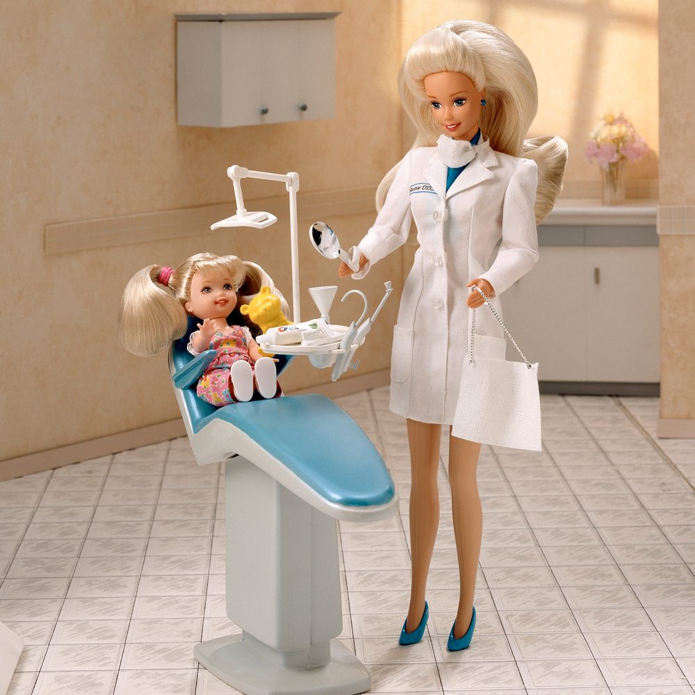 Doll dentist examines a seated child doll in a clinic setting with beige walls, floral decor, and medical tools.