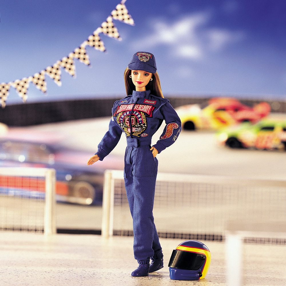 A doll wearing a blue racing jumpsuit stands on a racetrack with cars in the background; a helmet lies on the ground nearby.
