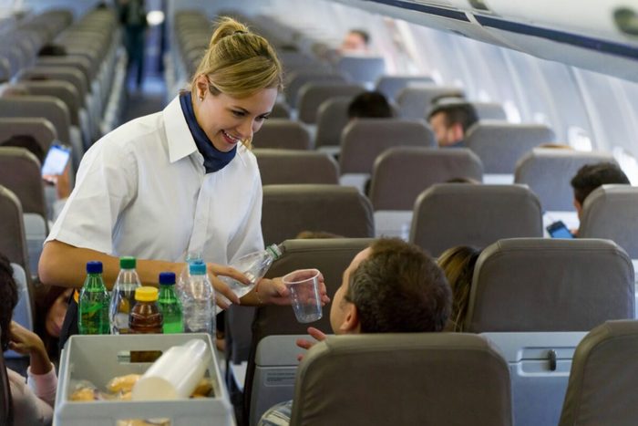 Flight attendant serves water to a passenger in an airplane aisle, with other passengers seated and occupied in a modern cabin environment.